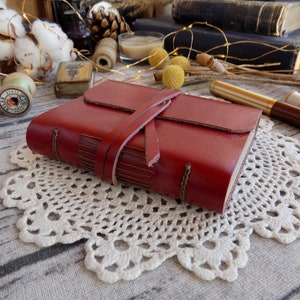 May include: A red leather-bound journal with a leather strap closure. The journal is sitting on a white crocheted doily.