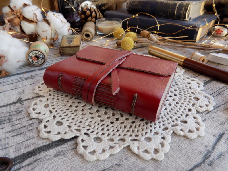May include: A red leather-bound journal with a leather strap closure. The journal is sitting on a white crocheted doily.