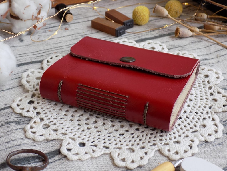 May include: A red leather-bound journal with a flap closure and a decorative stitch design. The journal is sitting on a white doily.
