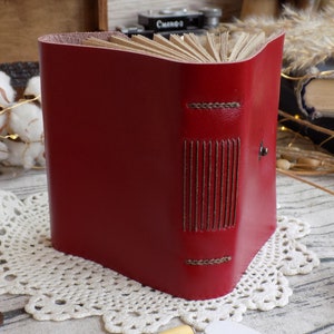 May include: A red leather-bound journal with decorative stitching and a metal clasp. The journal is closed and sitting on a white lace doily.