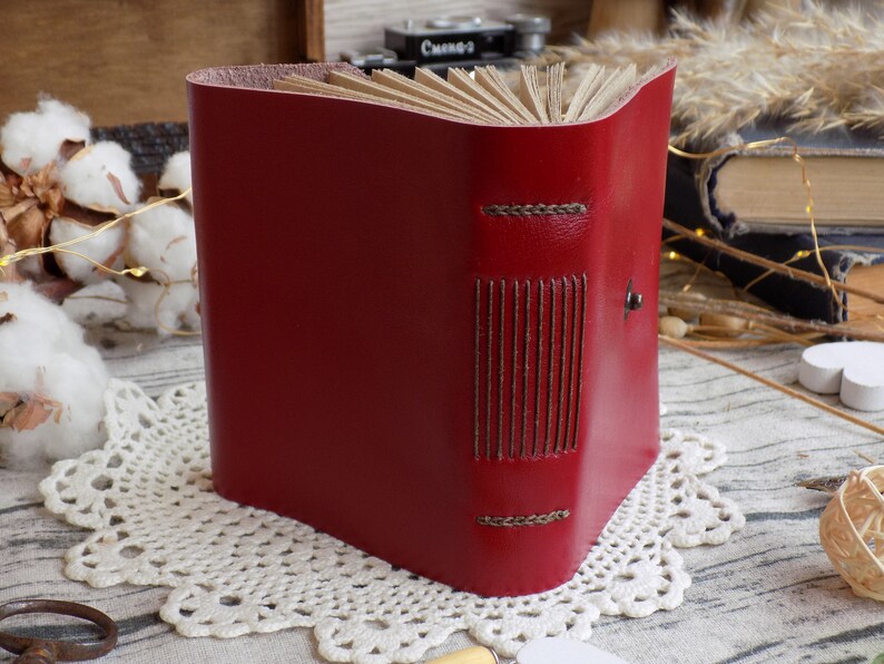 May include: A red leather-bound journal with decorative stitching and a metal clasp. The journal is closed and sitting on a white lace doily.