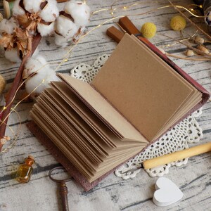 May include: A brown leather-bound journal with blank pages, open to reveal the pages. The journal is surrounded by decorative items, including cotton bolls, string, a small bottle, and a key.