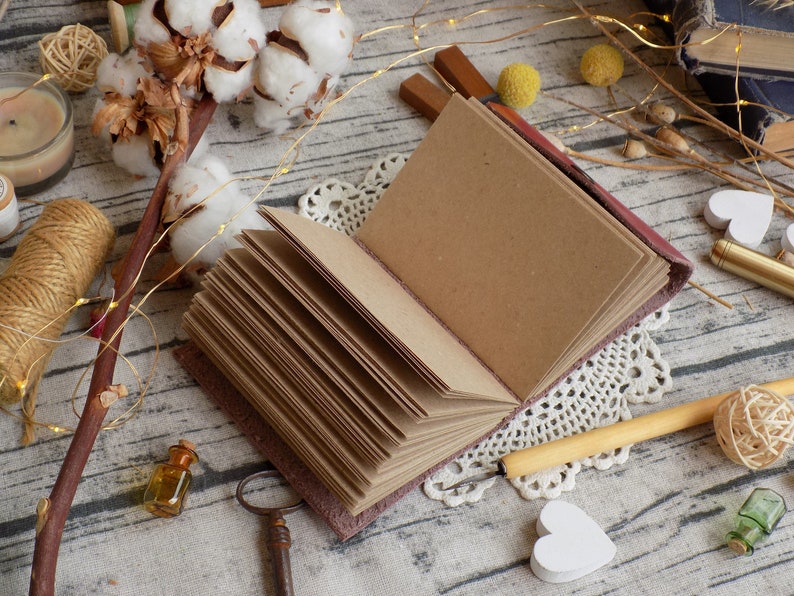 May include: A brown leather-bound journal with blank pages, open to reveal the pages. The journal is surrounded by decorative items, including cotton bolls, string, a small bottle, and a key.