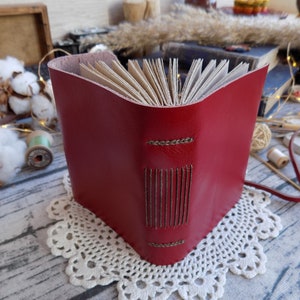 May include: A red leather-bound journal with a stitched spine and a brown leather strap. The journal is closed and sitting on a white doily.