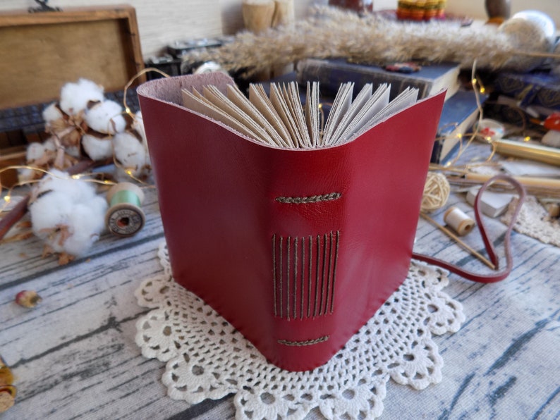 May include: A red leather-bound journal with a stitched spine and a brown leather strap. The journal is closed and sitting on a white doily.