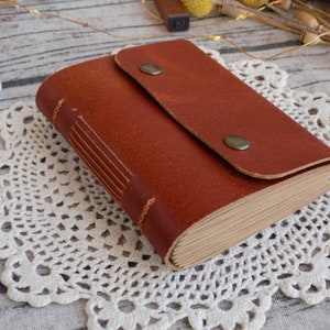 May include: A brown leather-bound journal with a flap closure and two metal button accents. The journal is lying on a white crocheted doily.