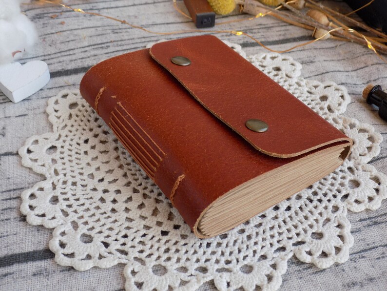 May include: A brown leather-bound journal with a flap closure and two metal button accents. The journal is lying on a white crocheted doily.