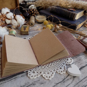 May include: A brown leather-bound journal with blank pages, open and lying on a white crocheted doily. The journal is surrounded by decorative items, including cotton bolls, a small wooden box, and a string of fairy lights.