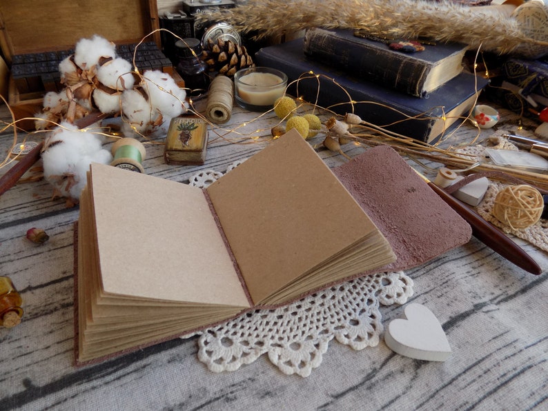 May include: A brown leather-bound journal with blank pages, open and lying on a white crocheted doily. The journal is surrounded by decorative items, including cotton bolls, a small wooden box, and a string of fairy lights.