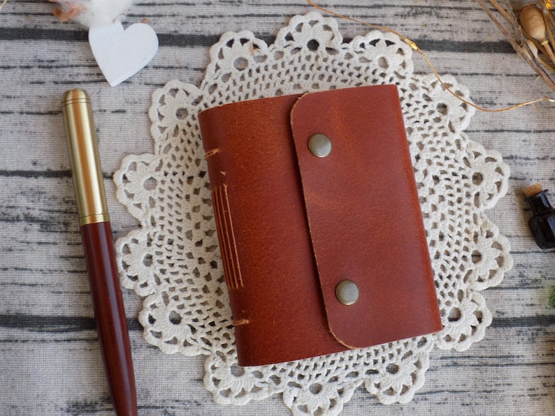 May include: A brown leather-bound journal with a flap closure and two brass clasps. The journal is sitting on a white doily.