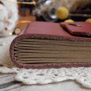 May include: A burgundy leather-bound journal with a thick stack of blank pages. The journal is closed and lying on a white crocheted doily.