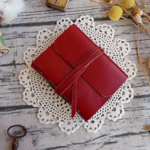 May include: A red leather journal with a tied closure, resting on a white doily. The journal is on a wooden surface with other decorative items.