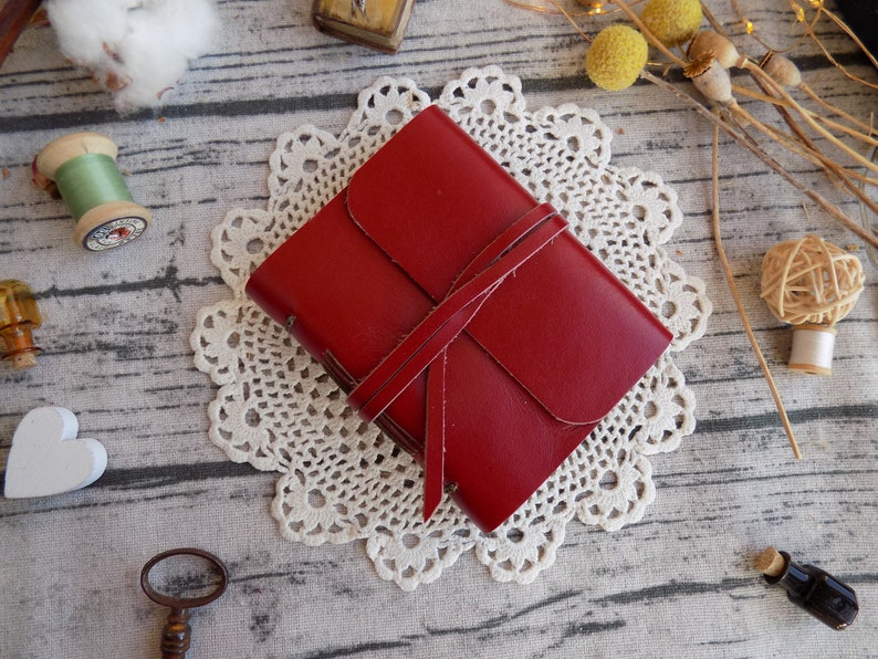 May include: A red leather journal with a tied closure, resting on a white doily. The journal is on a wooden surface with other decorative items.