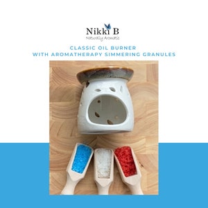 May include: White ceramic oil burner with a brown glaze on the top. The burner has a decorative cutout design. Three small wooden scoops filled with blue, white, and red granules are in front of the burner.