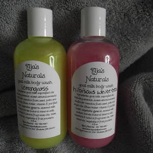 May include: Two clear plastic bottles of goat milk body wash. The bottle on the left is yellow and says "Aja's Naturals lemongrass". The bottle on the right is pink and says "Aja's Naturals hibiscus white tea".