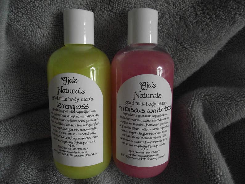 May include: Two clear plastic bottles of goat milk body wash. The bottle on the left is yellow and says "Aja's Naturals lemongrass". The bottle on the right is pink and says "Aja's Naturals hibiscus white tea".