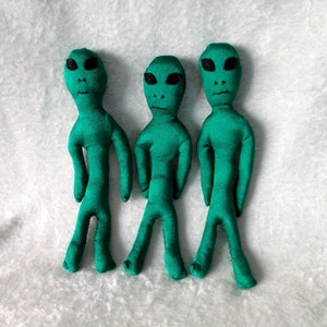 May include: Three green alien plush toys with black eyes and mouths. The aliens are standing upright on a white fuzzy surface.