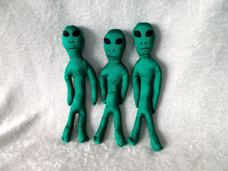 May include: Three green alien plush toys with black eyes and mouths. The aliens are standing upright on a white fuzzy surface.