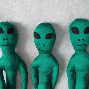 May include: Three green alien plush toys with black eyes and mouths. The aliens are standing upright on a white fuzzy surface.