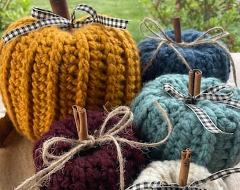 May include: Four crocheted pumpkins in different colours, including mustard yellow, teal, burgundy, and white, are arranged on a wooden tray with a white rim. Each pumpkin has a cinnamon stick stem and a black and white checked ribbon.