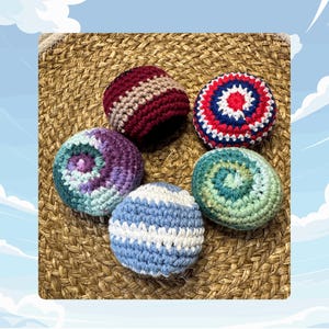 Puede incluir: Cinco bolas de ganchillo hechas a mano en varios colores, incluyendo rojo, azul, morado y verde. Las bolas están dispuestas sobre una superficie tejida marrón. Cada bola tiene un patrón y una combinación de colores únicos.