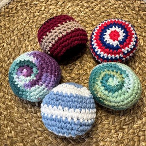 May include: Five colorful, crocheted balls in shades of red, blue, green, purple, and white. The balls are arranged on a woven, circular mat. Each ball features a unique pattern and color combination, showcasing handmade craftsmanship.