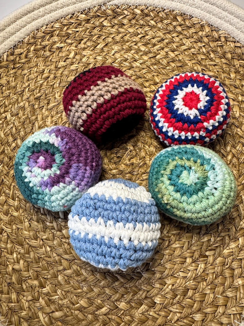 May include: Five colorful, crocheted balls in shades of red, blue, green, purple, and white. The balls are arranged on a woven, circular mat. Each ball features a unique pattern and color combination, showcasing handmade craftsmanship.