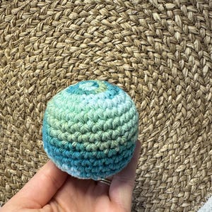 May include: A small, round, crocheted ball in shades of blue and green. The ball is held in a hand against a woven background. The yarn appears to be a variegated blend, creating a textured effect. The ball is approximately 2 inches in diameter.