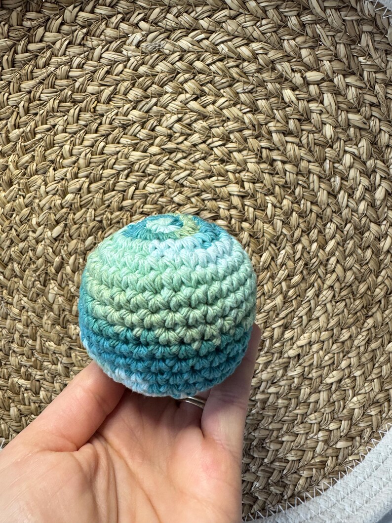 May include: A small, round, crocheted ball in shades of blue and green. The ball is held in a hand against a woven background. The yarn appears to be a variegated blend, creating a textured effect. The ball is approximately 2 inches in diameter.