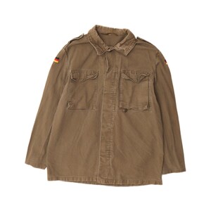 May include: An olive drab long-sleeve field jacket featuring a classic collar, two large button-flap chest pockets, and small German flag patches on both shoulders. The garment is constructed with a button-front closure and shoulder epaulets.