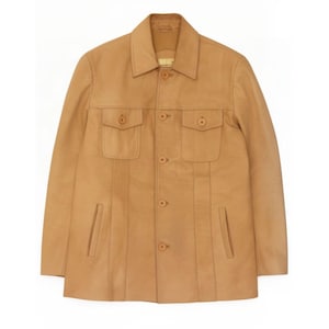 May include: A tan leather jacket featuring a classic collar, a central button-down front closure, and two chest pockets with buttoned flaps. The garment includes vertical seam detailing and two vertical slit pockets at the lower front section.