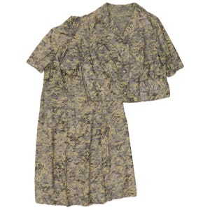 May include: A two-piece set consisting of a short-sleeved button-down shirt with a collar and two front pockets, and a matching knee-length skirt. Both pieces feature an abstract, mottled pattern in shades of muted olive green, grey, and black.