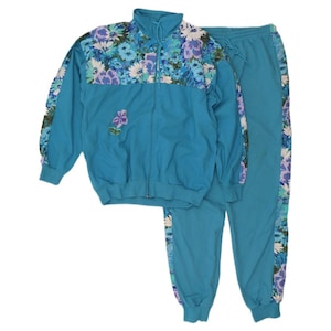 Puede incluir: Un chándal turquesa con una chaqueta con cremallera y pantalones a juego. La chaqueta tiene hombros con estampado floral y una pequeña flor bordada. Los pantalones tienen una franja floral en el lateral. El chándal está hecho de un material ligero.
