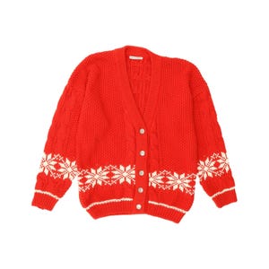 Könnte beinhalten: Eine leuchtend rote Strickjacke mit V-Ausschnitt, grobem Zopfmuster und einem weißen Schneeflockenmuster am unteren Rumpf und an den Ärmeln. Das Kleidungsstück verfügt über eine Knopfleiste mit fünf runden weißen Knöpfen und gerippte Bündchen.
