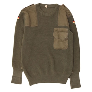 Może przedstawiać: Oliwkowy sweter w stylu wojskowym z wzmocnionymi naszywkami na ramionach i kieszenią na piersi. Posiada prążkowaną fakturę dzianiny i detal z flagą Niemiec na rękawie. Odzież w stylu vintage.