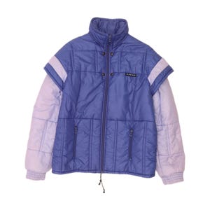 Può includere: Una giacca imbottita trapuntata nei toni del viola e del lavanda con collo alto, chiusura frontale con zip e due tasche laterali con zip. La giacca presenta un design a strati simile a un gilet sul busto e riporta la scritta "SADAS" su un'etichetta.