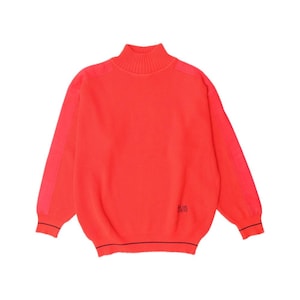 May include: Vibrant red sweater with a ribbed texture and a high neck. The sleeves feature a contrasting red panel. The hem and cuffs are trimmed with a thin black stripe. A small black logo is visible on the lower right side.