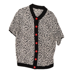 May include: A short-sleeved cardigan featuring a black and white floral pattern throughout. The garment includes a solid black V-neck collar, a black button placket, and a black hem. Seven bright red buttons are aligned vertically down the front.