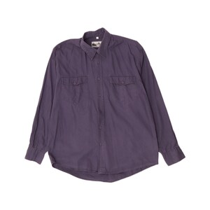 May include: A long-sleeve button-down shirt in a muted dark purple color. The shirt features a standard collar, a central button placket, and two chest pockets with buttoned flaps. A small white rectangular label is visible at the inner neckline.