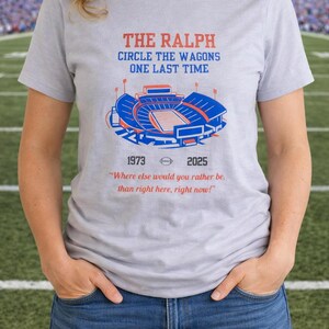 Woman wearing a gray Buffalo Bills shirt featuring “The Ralph – Circle the Wagons One Last Time” stadium graphic with 1973–2025 tribute, standing on a football field background.