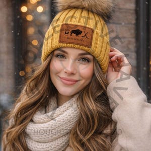 May include: Mustard yellow knit beanie with a large faux fur pom-pom. The beanie features a brown leather patch with a buffalo and hockey stick graphic, and the text "BUFFALO, NY". A cream-colored scarf and tan coat complete the winter look.