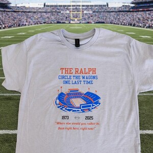 Flat lay of gray Buffalo Bills shirt featuring “The Ralph – Circle the Wagons One Last Time” stadium graphic with 1973–2025 tribute, displayed on a football field background.