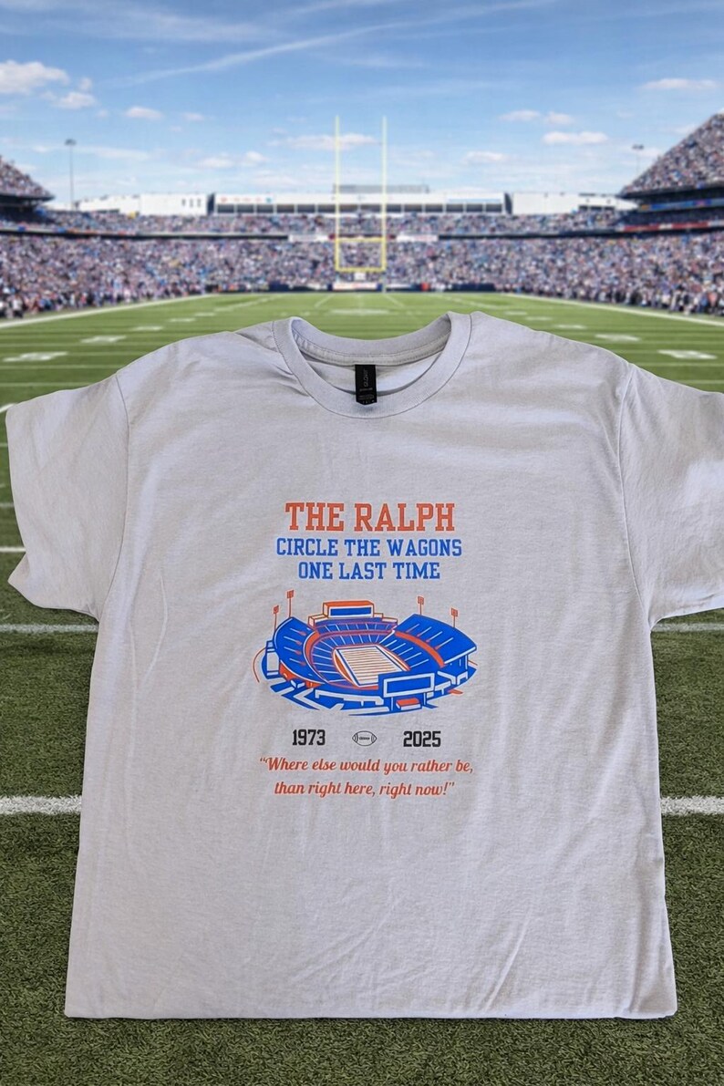 Flat lay of gray Buffalo Bills shirt featuring “The Ralph – Circle the Wagons One Last Time” stadium graphic with 1973–2025 tribute, displayed on a football field background.
