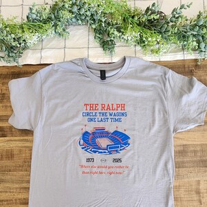 Flat lay of gray Buffalo Bills shirt with “The Ralph – Circle the Wagons One Last Time” stadium design, styled on a wooden table with greenery and neutral décor.