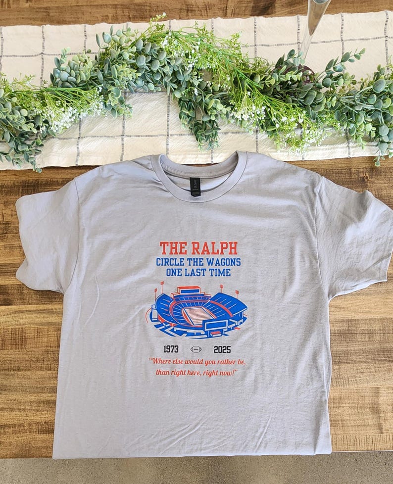 Flat lay of gray Buffalo Bills shirt with “The Ralph – Circle the Wagons One Last Time” stadium design, styled on a wooden table with greenery and neutral décor.
