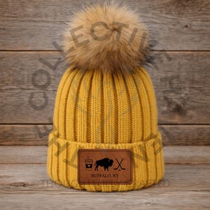 May include: Mustard yellow knit beanie with a large faux fur pom-pom. A brown leather patch features a coffee cup, buffalo, hockey stick, and the text "BUFFALO, NY". The hat is made of a ribbed knit material.