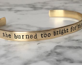 May include: A gold-toned metal cuff bracelet with an open back design. The exterior surface features the stamped black text "she burned too bright for this world" in a simple, lowercase sans-serif font, following the curve of the band.