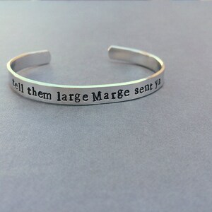 May include: A silver-coloured metal cuff bracelet with an open back. The exterior surface features the stamped black text "tell them large Marge sent ya" in a typewriter-style font, running horizontally along the length of the band.