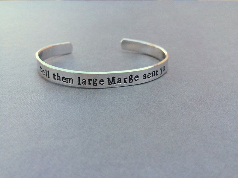 May include: A silver-coloured metal cuff bracelet with an open back. The exterior surface features the stamped black text "tell them large Marge sent ya" in a typewriter-style font, running horizontally along the length of the band.