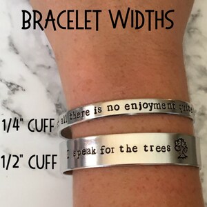 May include: Two silver metal bracelets with engraved text. The top bracelet is 1/4 inch wide and reads "& all there is no enjoyment quite". The bottom bracelet is 1/2 inch wide and reads "I speak for the trees" with a small tree symbol.